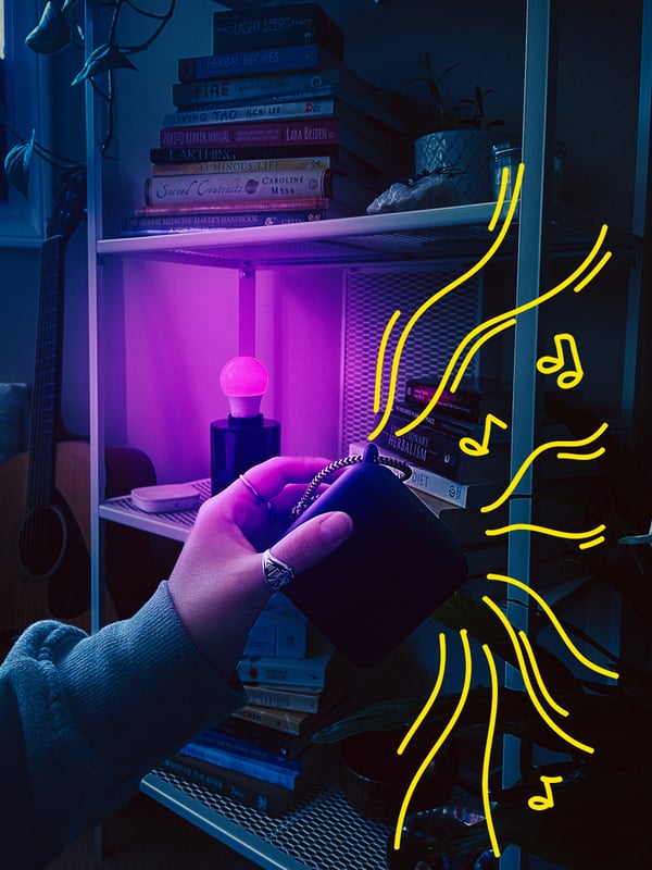 A hand holding a VAPPEBY bluetooth speaker, with a TRÅDFRI light bulb shining a purple-coloured light in the background.