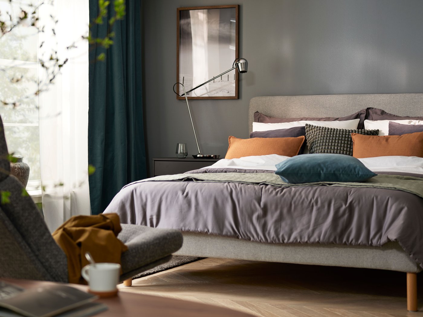 Recámara con paredes grises y vista de una cama, un buró y estantes. En primer plano, hojas verdes difuminadas de una planta.