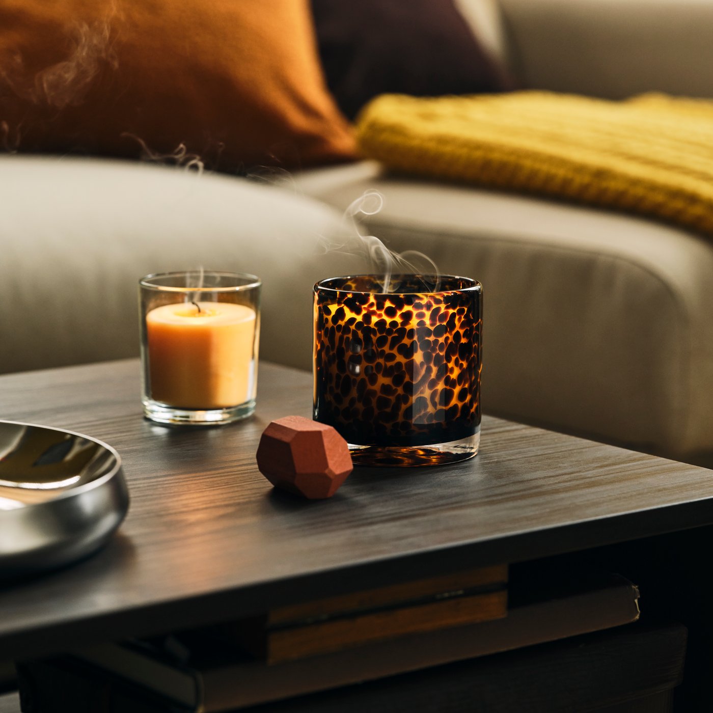 A brown KRONSÅNGARE scented candle in a dotted glass holder next to another candle, both placed on a HOLMERUD side table.