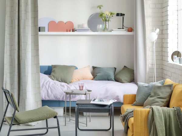 A pastel 24-hour living room oasis with a medium blue bed, a yellow FRIDHULT sofa-bed and an olive-green easy chair.