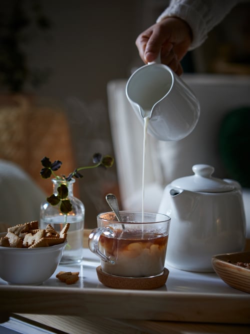 Gros plan sur une personne versant le lait d’un pot à lait dans une tasse IKEA 365+ en verre transparent remplie de thé.