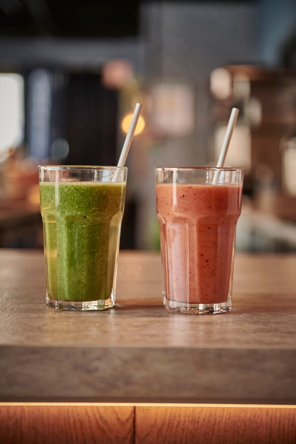 Två smoothies i glas, en röd och en grön, båda med vita sugrör. De står sida vid sida på en grå bänkskiva.