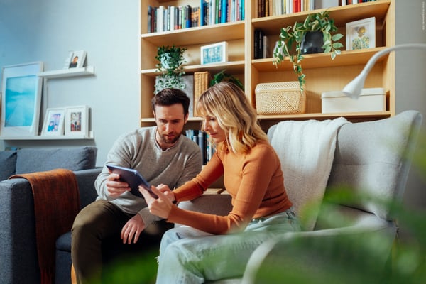 A couple is sitting on a green sofa in their living room and are looking at their tablet