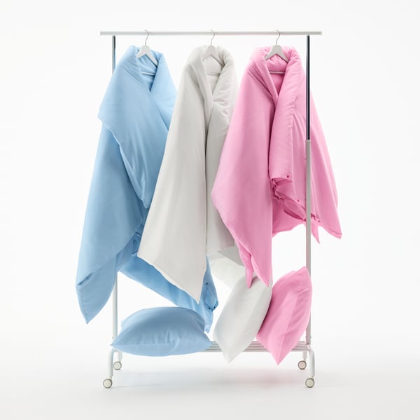 Three PILTANDVINGE duvet sets in light blue, white and pink hang on a clothing rack. On a shelf below, matching pillows.