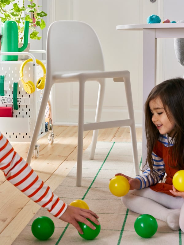 An adult and a child play with some balls on a rug near an URBAN junior chair and a BRÄNNBOLL shelving unit on castors.
