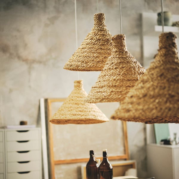 Närbild på fyra konformade lampskärmar i naturmaterial, hängande i ett rustikt rum med vita lådor och bruna flaskor.
