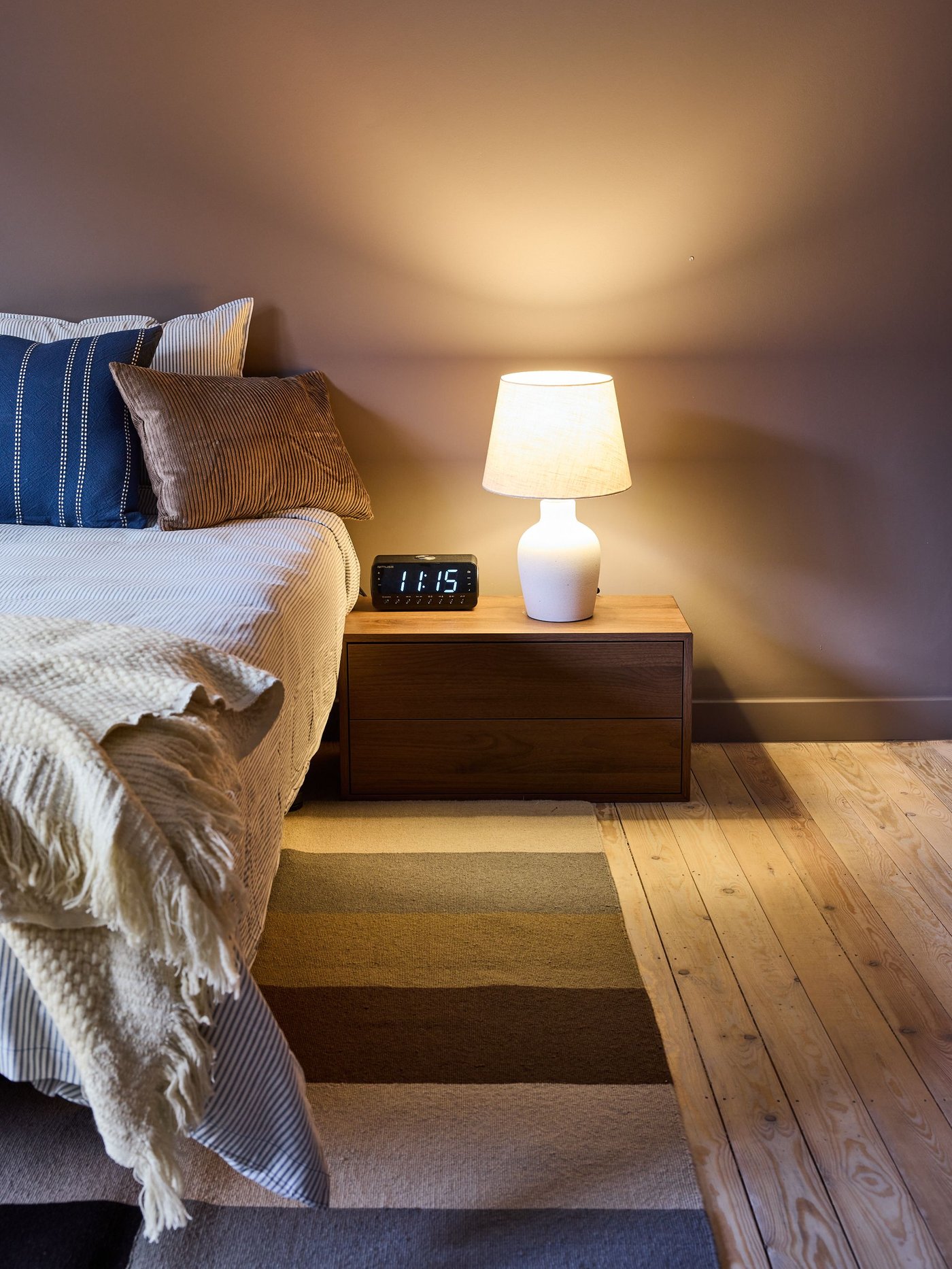 Slaapkamer met bed en kussens naast houten nachtkastje met lamp en wekker, warme verlichting, tapijt en houten vloer in rustige sfeer.
