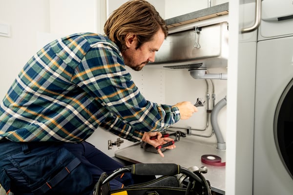 Un tizio in tuta da lavoro sta stringendo con delle pinze i raccordi dell'acqua sotto un lavandino.