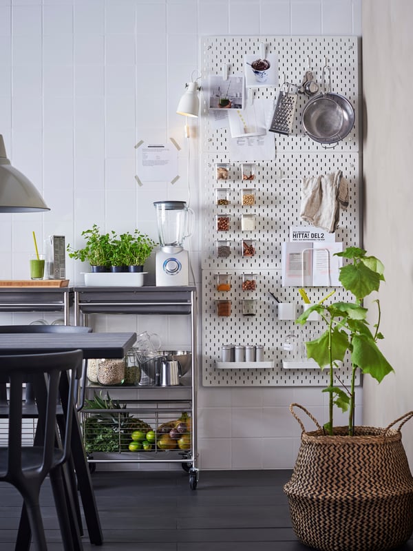 Hvide SKÅDIS opbevaringstavler er monteret på en køkkenvæg ved siden af et køkkenbord med tilbehør til opskrifter og ingredienser til smoothies.