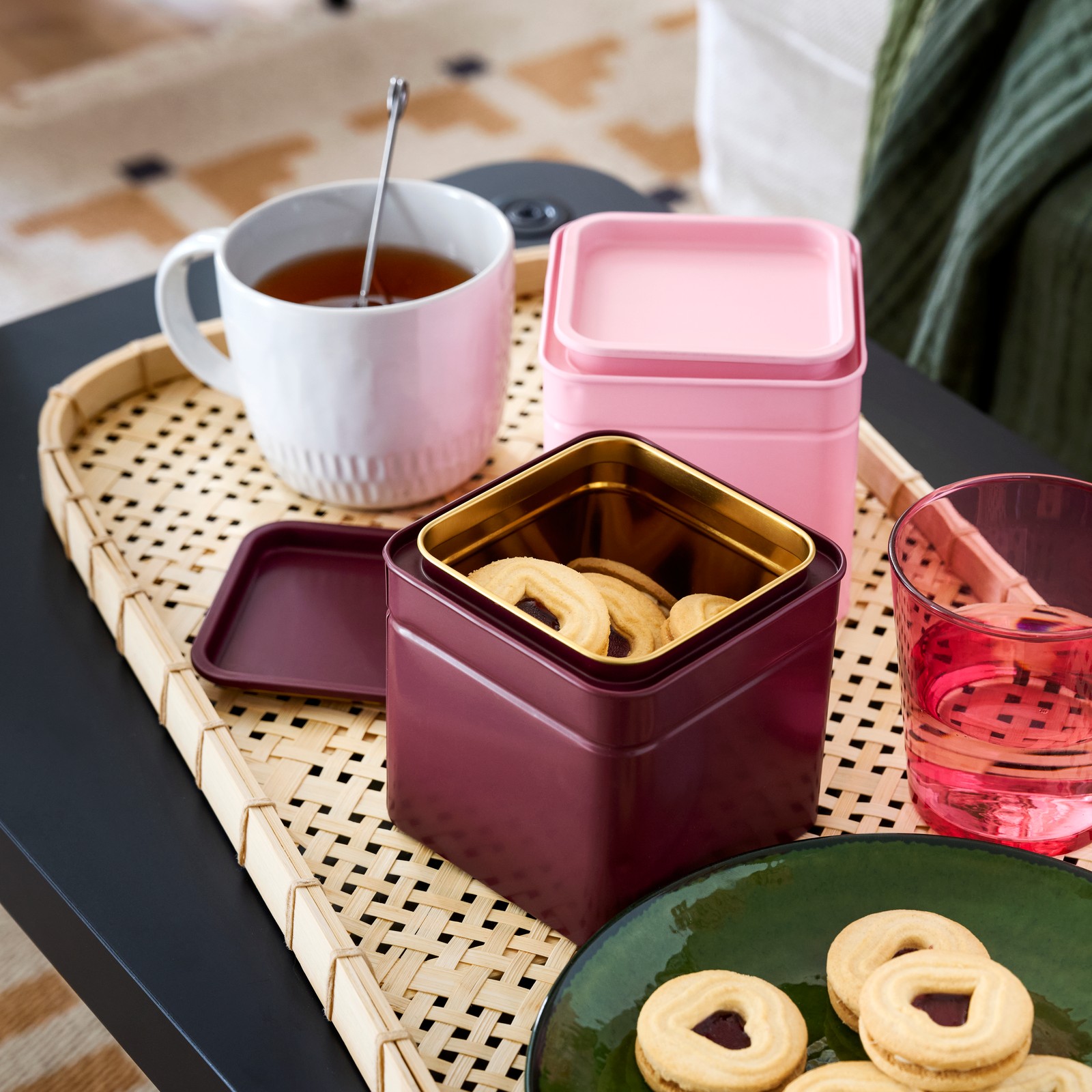 A BLOMNING tin with lid in dark red, shown open with cookies inside, placed on tray with tea, glasses and biscuits.
