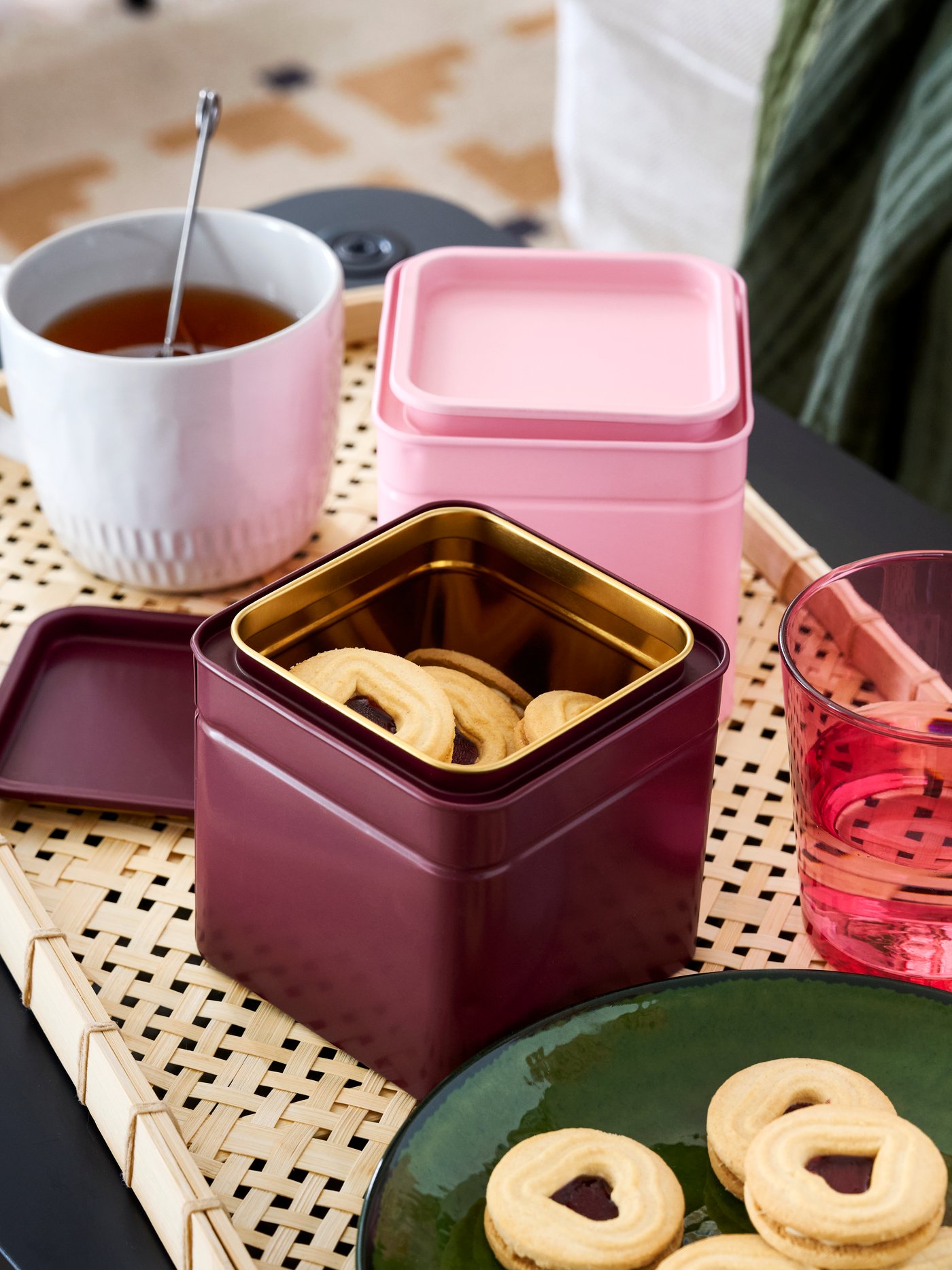 A BLOMNING tin with lid in dark red, shown open with cookies inside, placed on tray with tea, glasses and biscuits.