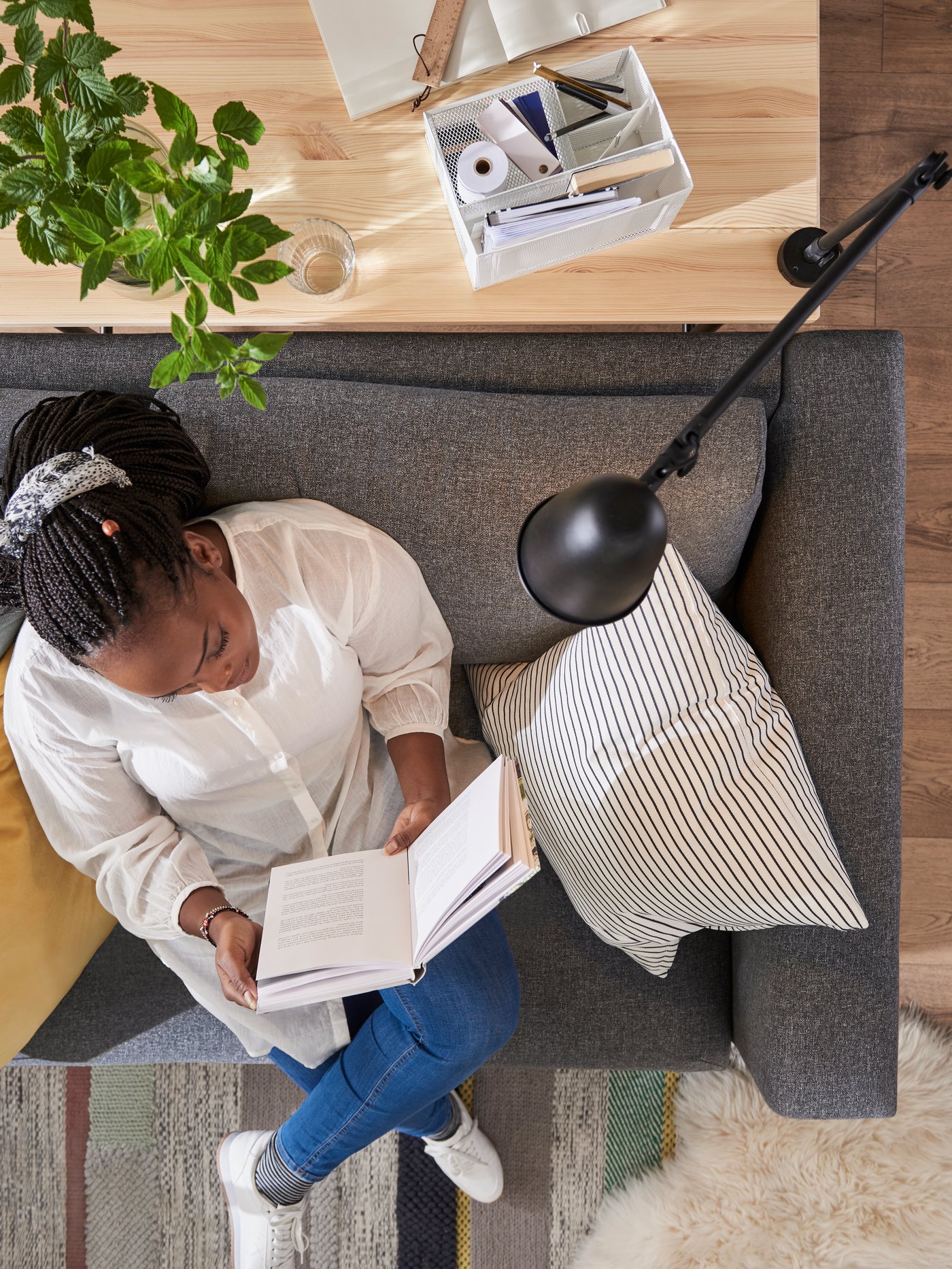 A woman reads a book on a VIMLE sofa-bed, under a SKURUP work/wall lamp attached to the KULLABERG desk behind her.