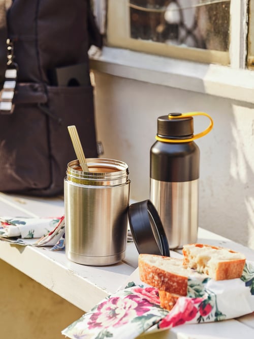 A spoon resting in an opened EFTERFRÅGAD food vacuum flask, placed next to an HETLEVRAD insulated flask on a white bench.