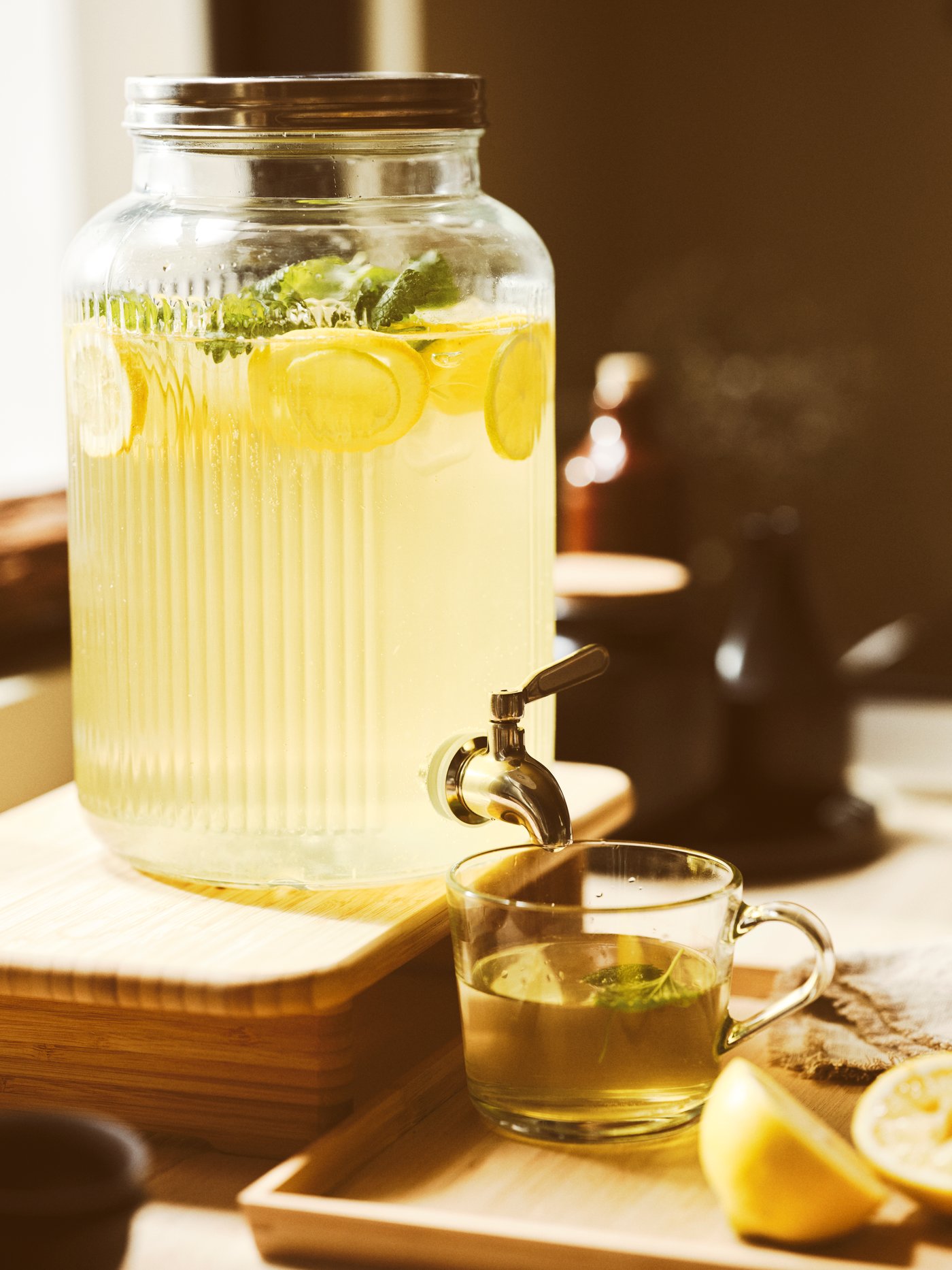 Una jarra de cristal VARDAGEN con grifo que contiene limonada está sobre una tabla de cortar STOLTHET. Debajo del grifo de la jarra hay una taza con líquido.