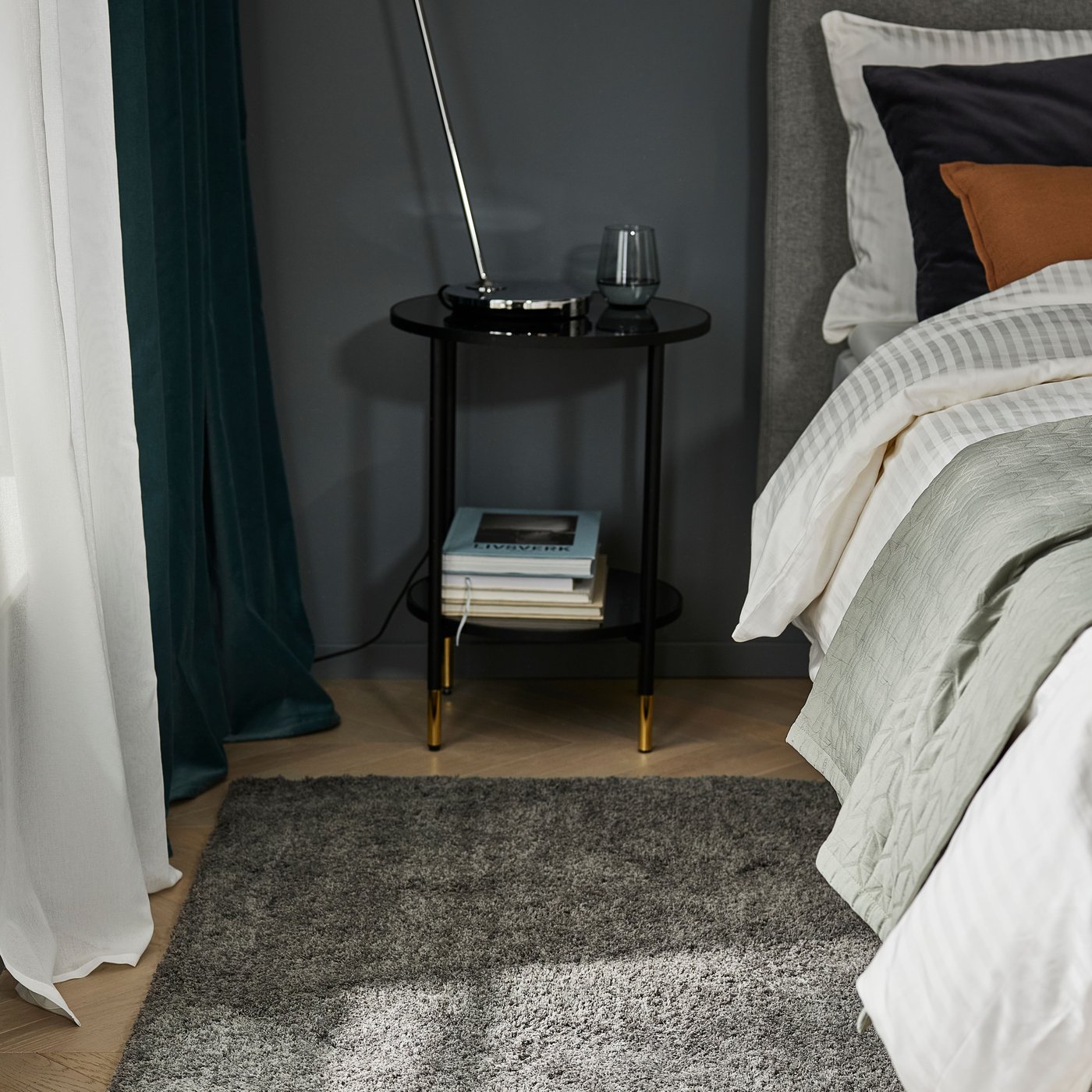 Ett modernt sovrum med ett svart sidobord i två nivåer bredvid en prydligt bäddad säng. En grå ryamatta ligger på trägolvet framför bordet, som har en lampa, ett glas och böcker.