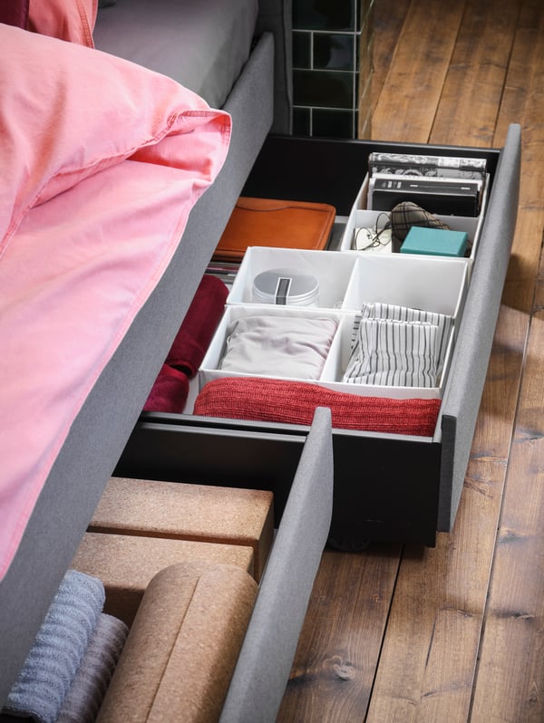 Two, grey, HAUGA upholstered bed storage boxes partly pulled out from beneath a grey, HAUGA bed, various small items inside.