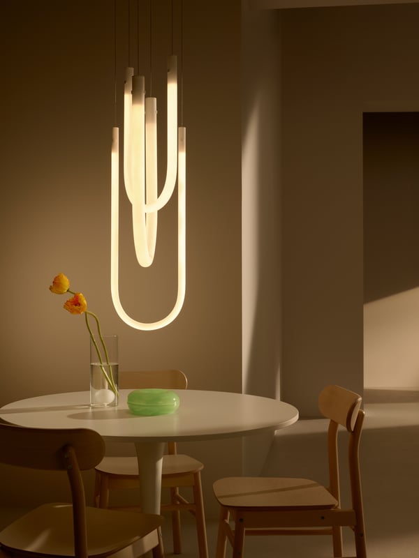 Una lampada a sospensione VARMBLIXT LED è sospesa sopra un tavolo rotondo con tre sedie e un vaso.