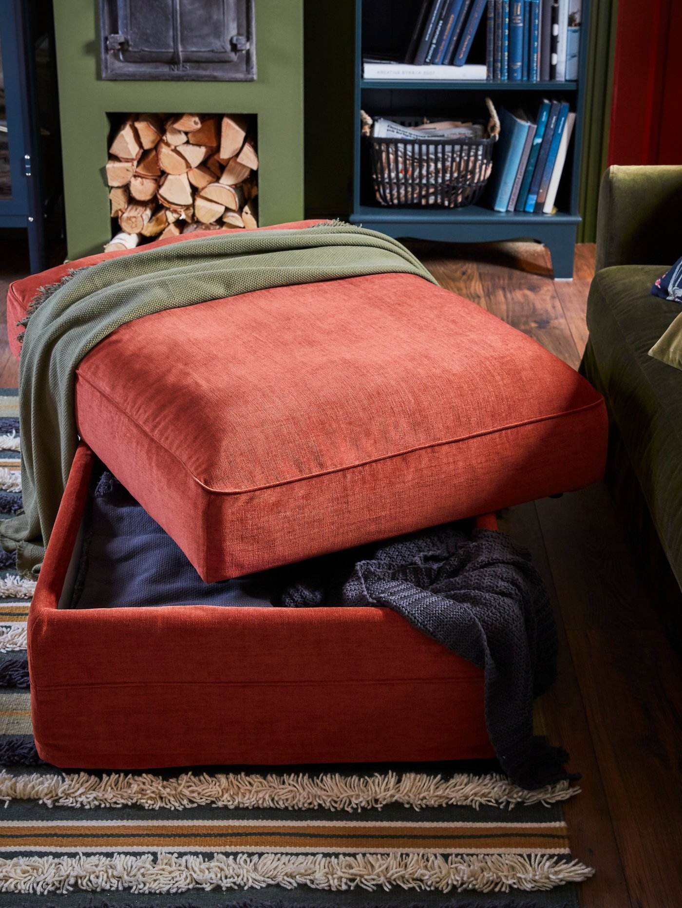 A light red footstool where the seat is open, throws are stored inside, with a dark blue-green bookcase behind.