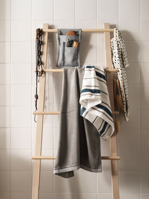 Badezimmer mit weiß gefliester Wand, davor steht ein VILTO Handtuchständer mit verschiedenen Handtüchern und einem Hängeutensil. Seitlich sind Schmuck und ein Babystrampler befestigt.