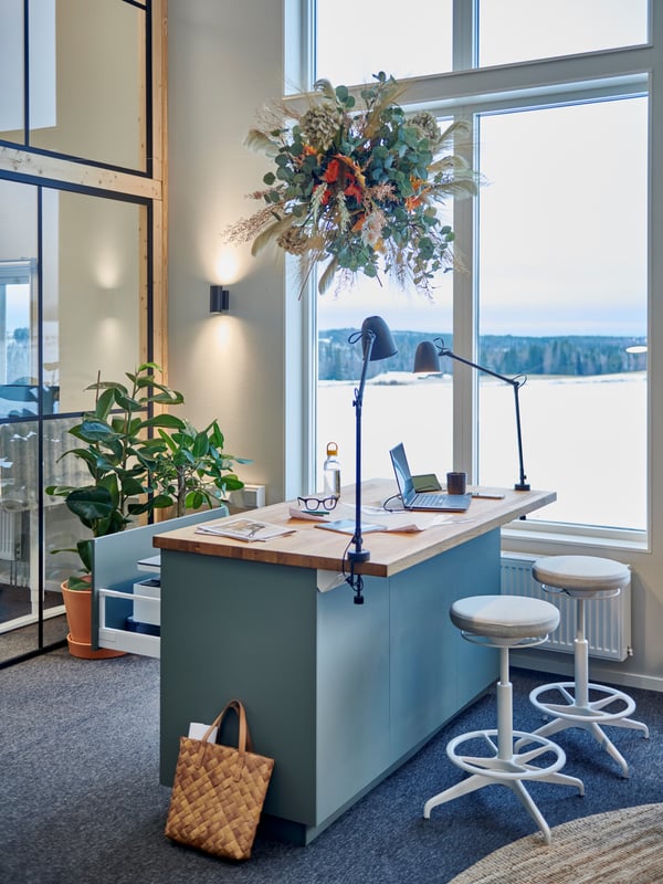 Modernt kontorsutrymme med stort fönster mot ett utomhuslandskap. Ett skrivbord med träskiva, två vita pallar, laptop, glasögon, papper och en skrivbordslampa. Hängande växt ovanför skrivbordet, grön krukväxt på golvet, hyllor med förvaringslådor och en flätad väska bredvid skrivbordet.