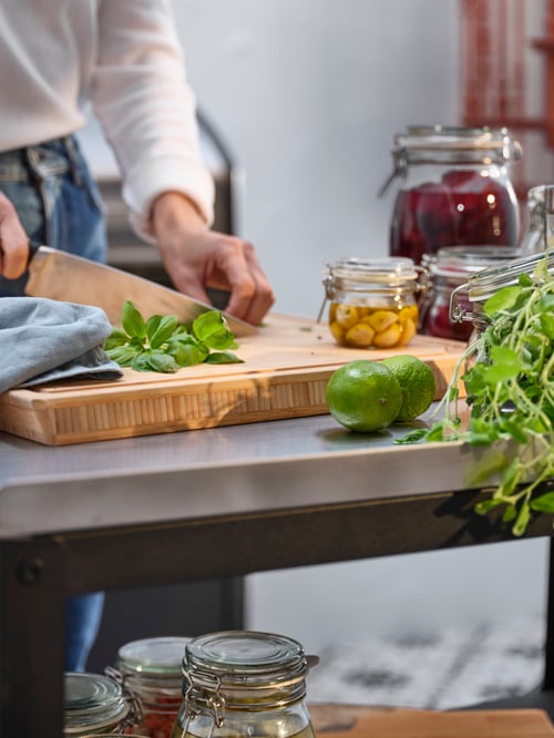 O femeie toacă busuioc proaspăt pe un tocător de carne APTITLIG. În apropiere, trei borcane KORKEN pline cu legume conservate.