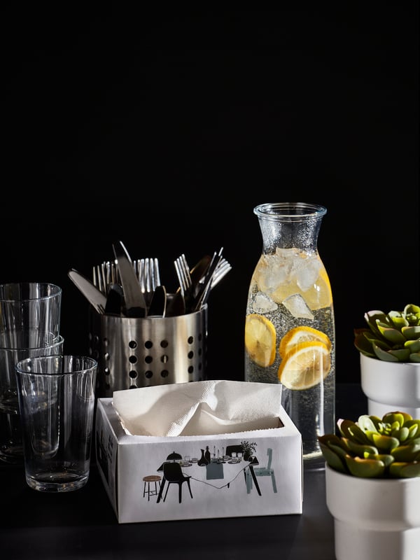 Display of glasses, cutlery, a carafe with water and slices of lemon, white paper napkins and two small plants.