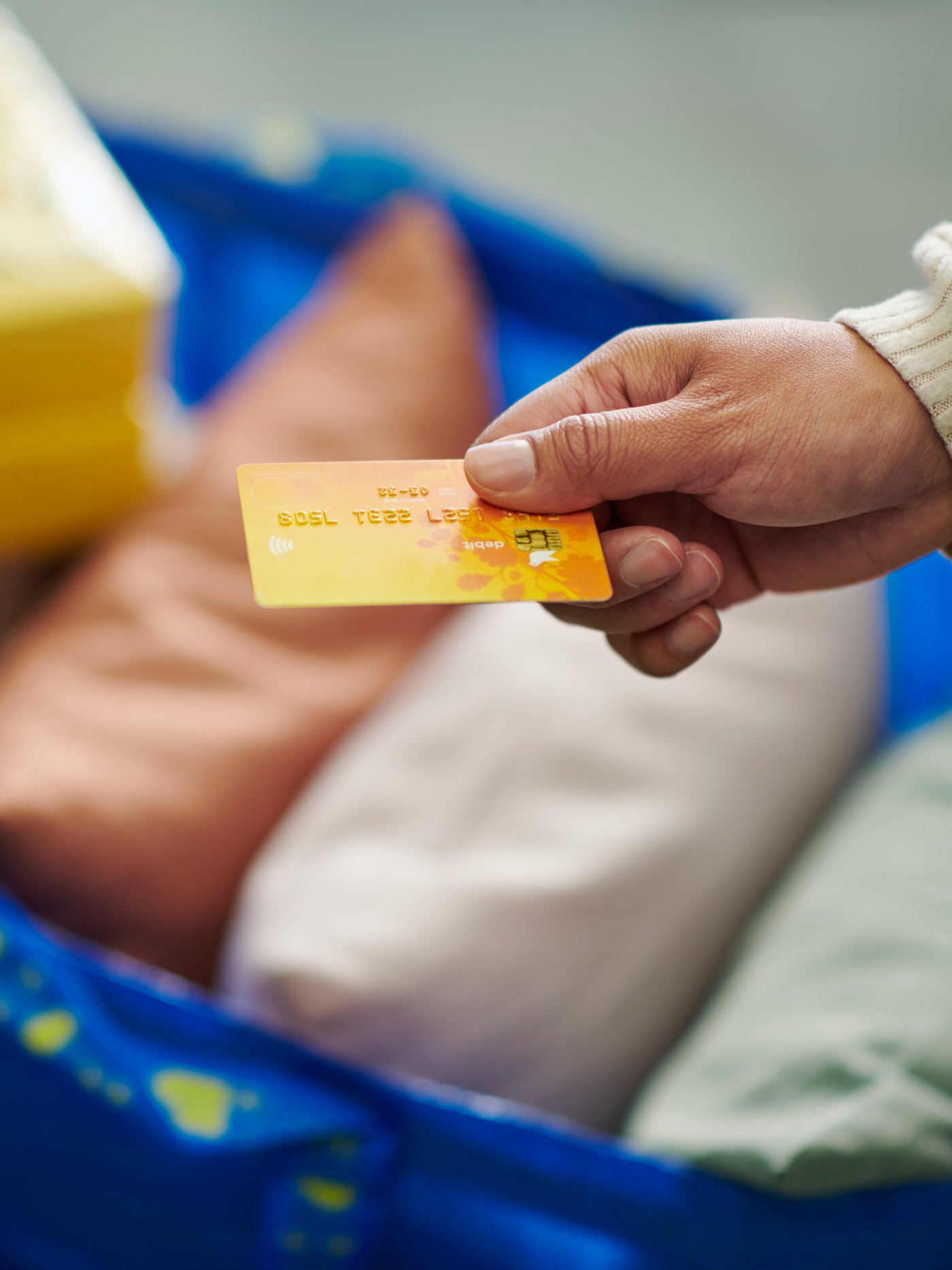 L’image montre une main tendant une carte bancaire orange, prête à effectuer un paiement. En arrière-plan, un grand sac bleu IKEA rempli d’articles textiles (coussins colorés) et de boîtes jaunes suggère une séance de shopping. La scène illustre un moment d’achat simple, pratique et quotidien, dans un univers IKEA reconnaissable.