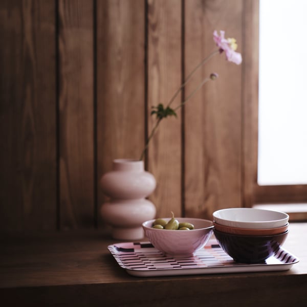 Primer plano de tazones HÖSTAGILLE en colores rosa, café, blanco roto y naranja, colocados sobre una charola estampada junto a un florero rosa en el alféizar de una ventana.