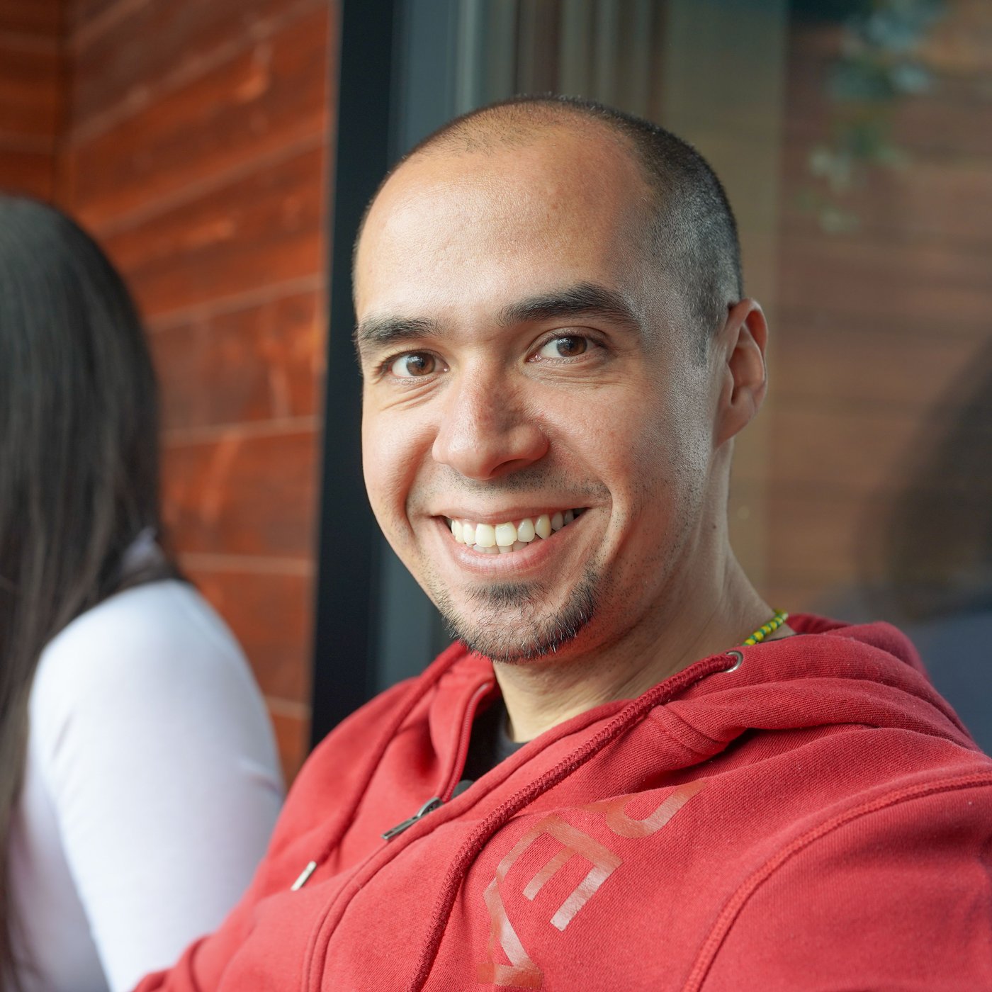 A man sits outdoors in his red shirt and smiles at the camera.