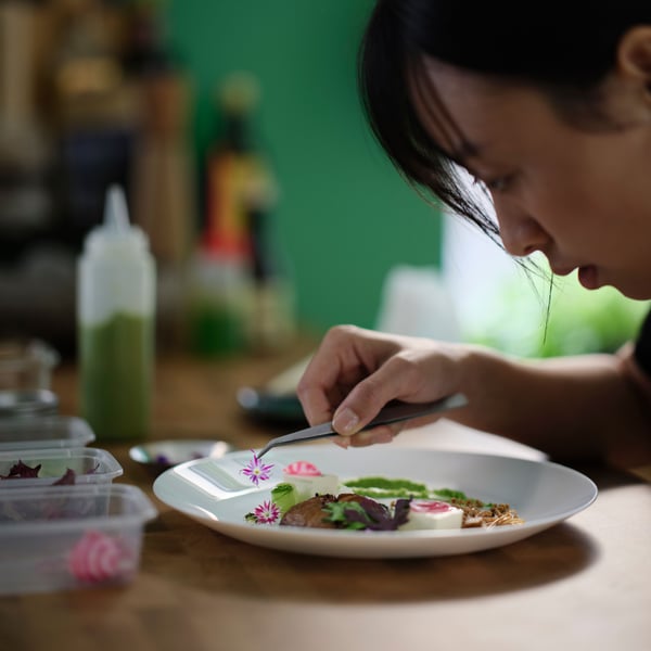A person is leaning over a white OFTAST deep plate with food on it, and carefully places a flower on the food using tweezers.