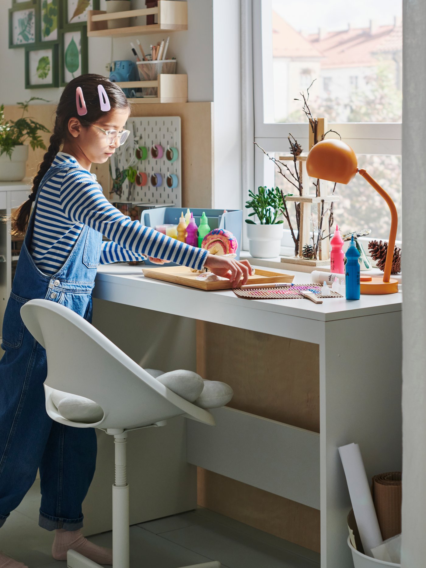 A young girl stands at a white LASTARE desk while lifting an OSTBIT bamboo tray featuring her latest hobby project: a snail.