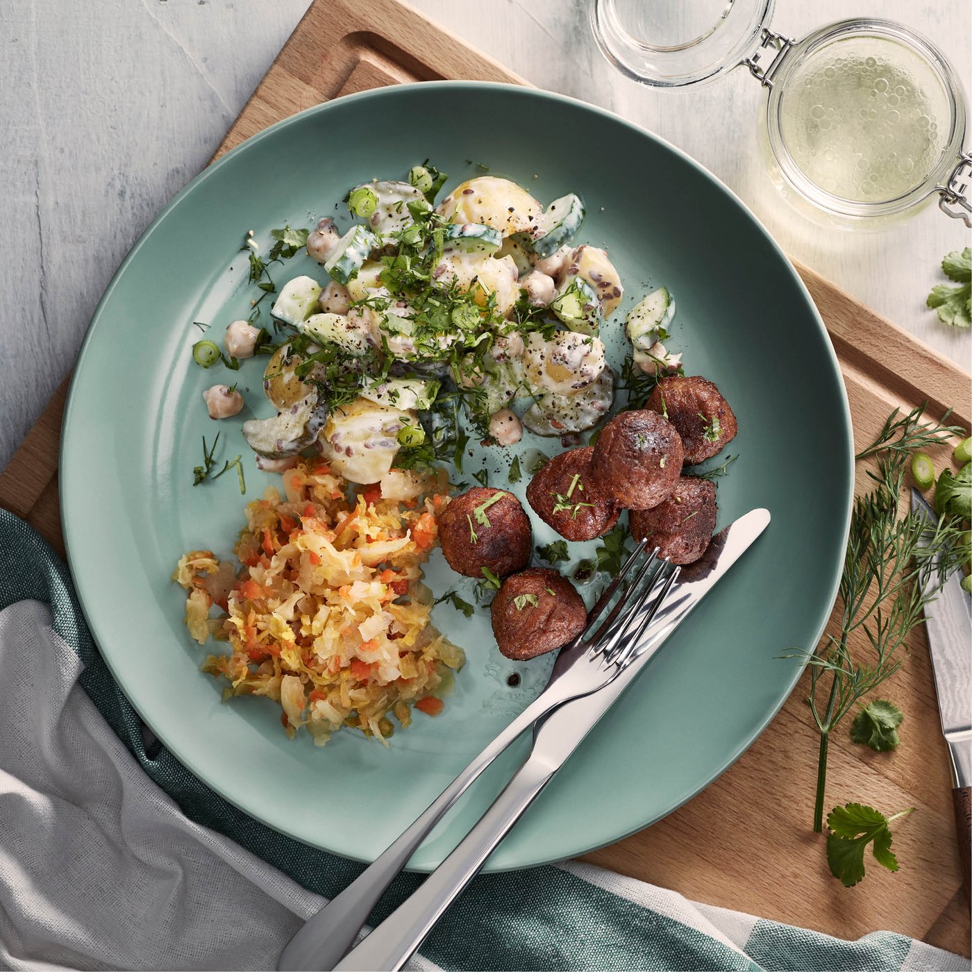 Bord met aardappelsalade, gegrilde worstschijfjes en gestoofde groenten, gegarneerd met verse kruiden op een houten tafel.