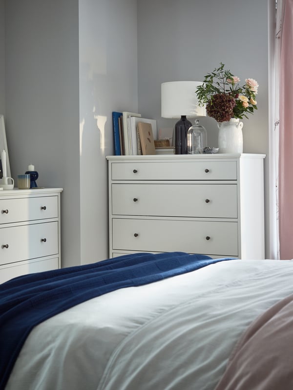 Two white chests of drawers in a corner with books, a lamp, a white vase with flowers, and candles on top.