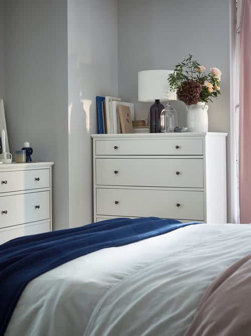 Two white chests of drawers in a corner with books, a lamp, a white vase with flowers, and candles on top.