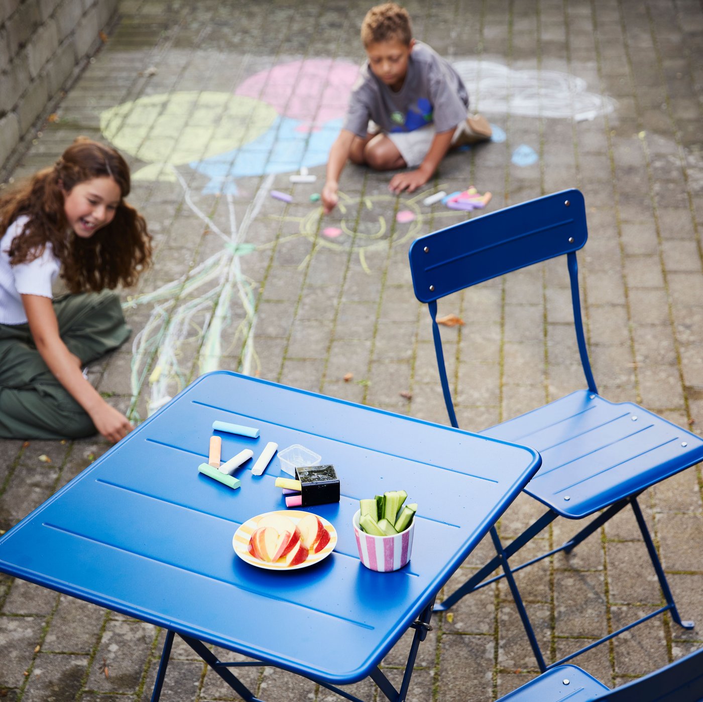 A blue SUNDSÖ table for two in a courtyard, with two kids in the background drawing on the pavers using MÅLA chalks.