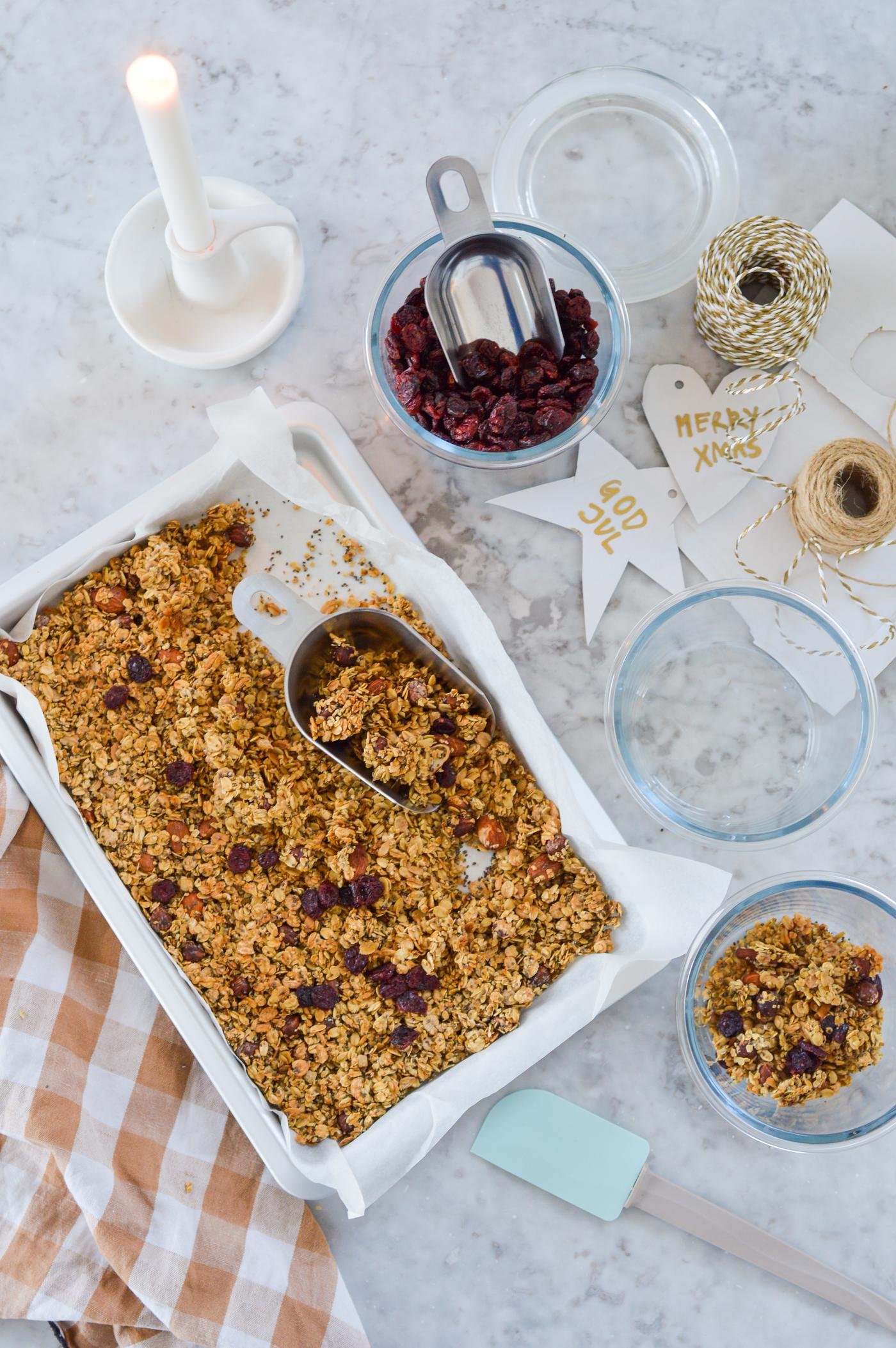 Müsli in einer weissen Auflaufform auf einer Marmoroberfläche, umgeben von einer brennenden Kerze, einem Glas Cranberries, Schnur, Weihnachtsdekoration und einem Küchentuch.