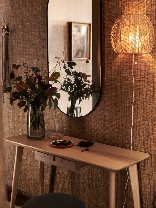 Ein ovaler LINDBYN Spiegel hängt über einem LISABO Schreibtisch mit einer Blumenvase und anderen Gegenständen. Daneben befindet sich eine MÅNALG Wandleuchte.