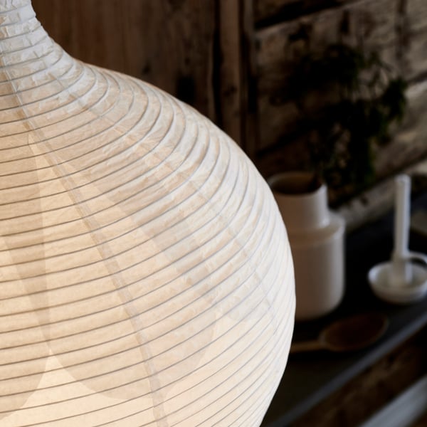 Primo piano di una lampada RISBYN, lo sfondo è sfocato ma si nota una mensola nera con un vaso bianco e alcune decorazioni.