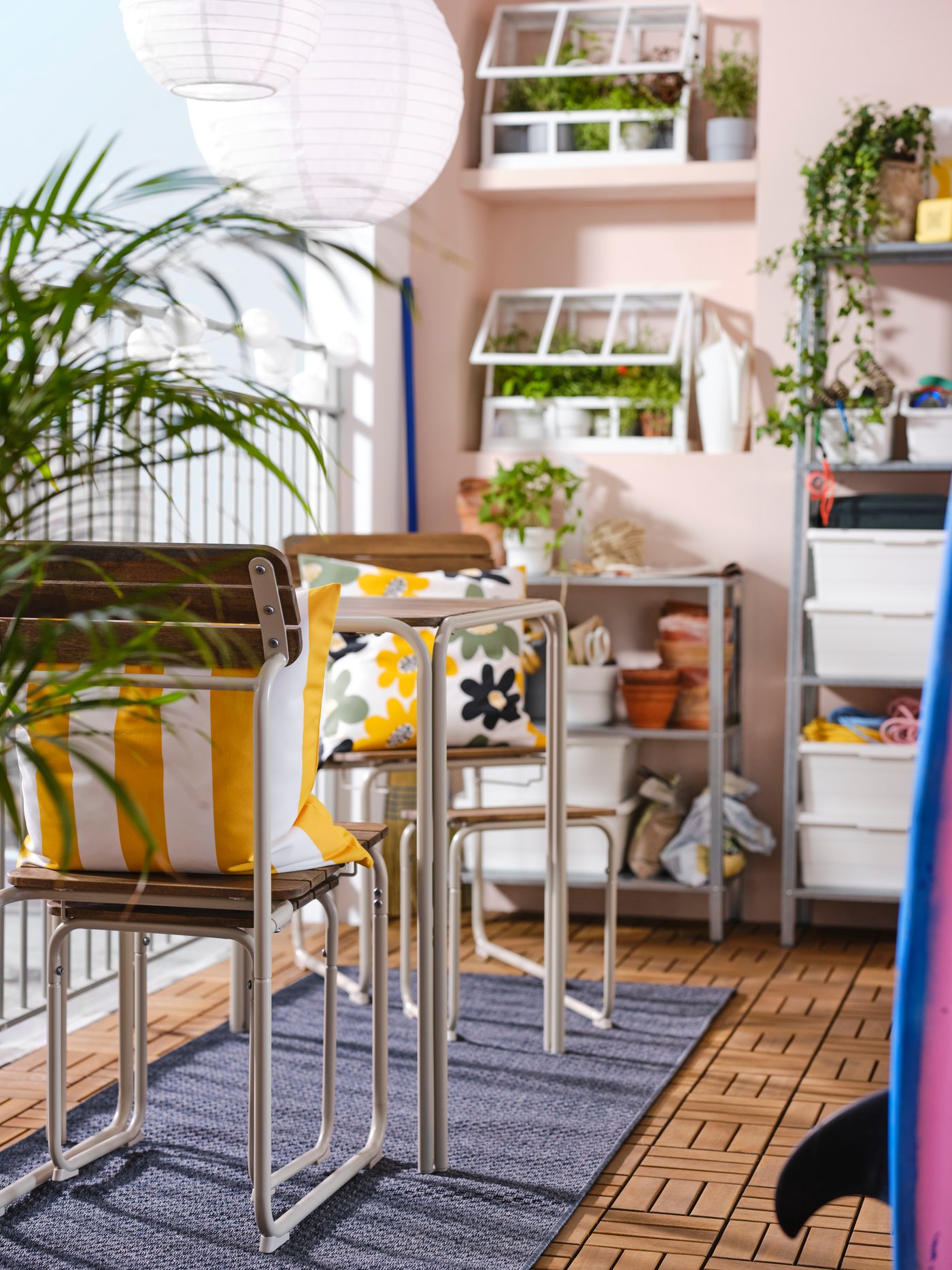 A balcony with pink walls featuring a FURUÖN dining table with 2 chairs and 2 footstools, shelving units and a greenhouse.