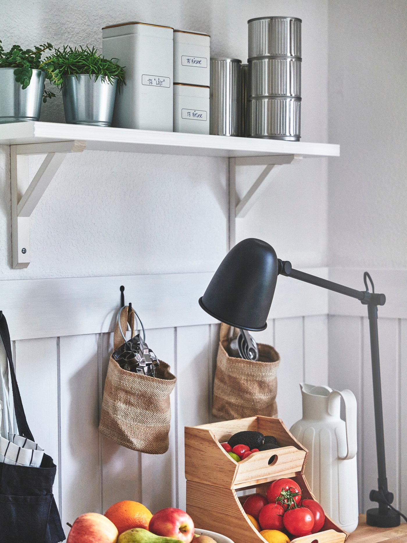 Träbord med en trälåda fylld med frukt framför en bordslampa med lång, justerbar arm. På bordet står även en vit kanna. Ovanför bordet finns en hylla med burkar och två hängande korgar.