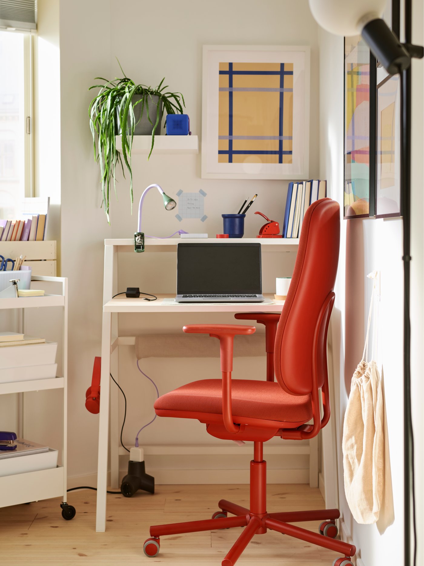 A small corner workspace with a MITTPLAN desk, a red SMÖRKULL office chair, a laptop, and a white trolley with storage boxes.