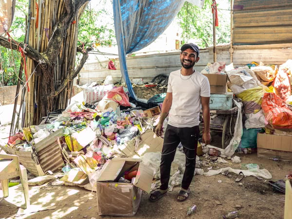 A person stands in a cluttered area filled with an assortment of discarded items. There are piles of plastic bottles, containers, and bags, alongside cardboard boxes and general refuse. The setting is outdoors with trees visible in the background. The person wear a white t-shirt, dark pants, and sandals.