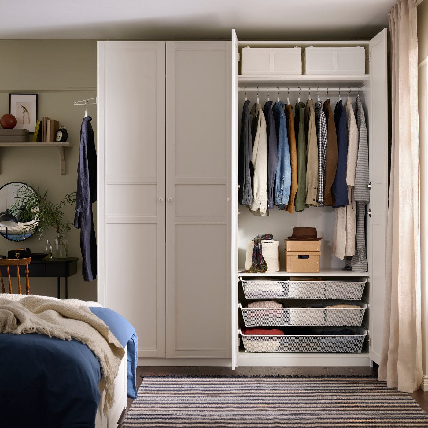 A bedroom with an open white PAX wardrobe with clothes hanging inside and KOMPLEMENT mesh baskets at the bottom.
