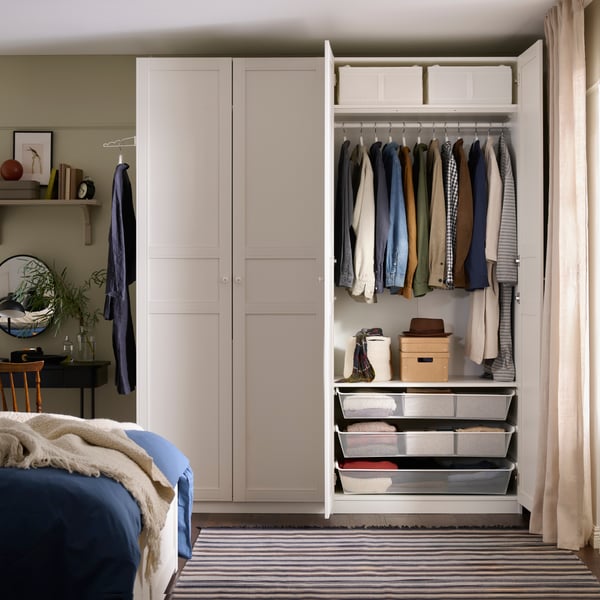 A bedroom with an open white PAX wardrobe with clothes hanging inside and KOMPLEMENT mesh baskets at the bottom.