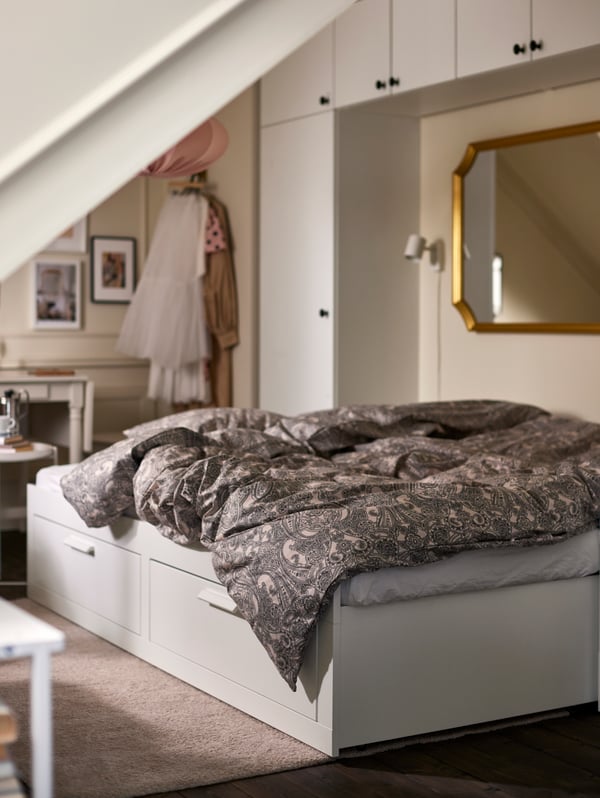 An extended white BRIMNES daybed made with beige/dark grey bedlinen, with white wardrobes and a gold-frame mirror behind.