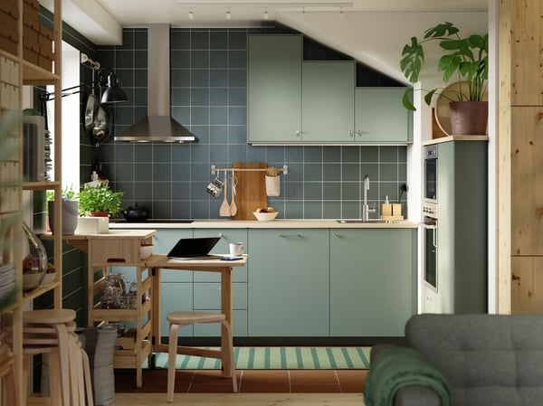 Eine kleine Küche mit matten, grau-grünen NICKEBO-Fronten hat Hängeschränke, die der Schräge der Decke folgen, und einen Holztisch.