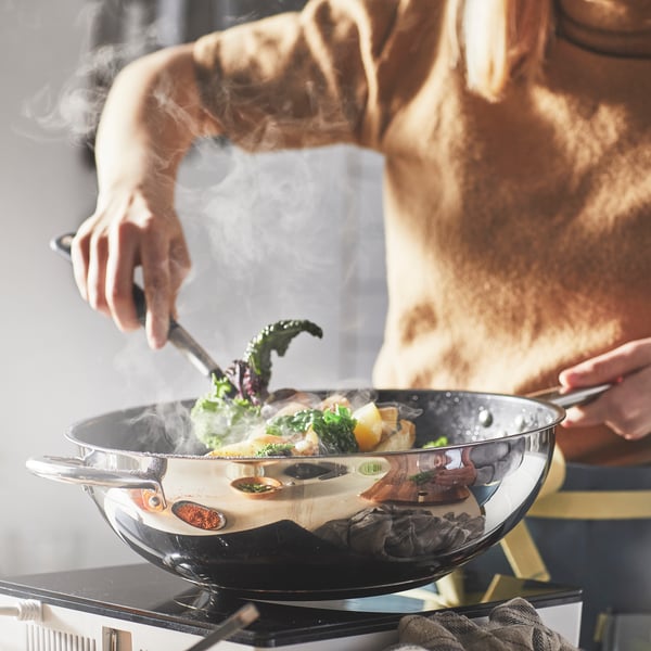A person in a brown apron is holding an IKEA 365+ wok of stainless steel and non-stick coating, stirring a mix of vegetables.