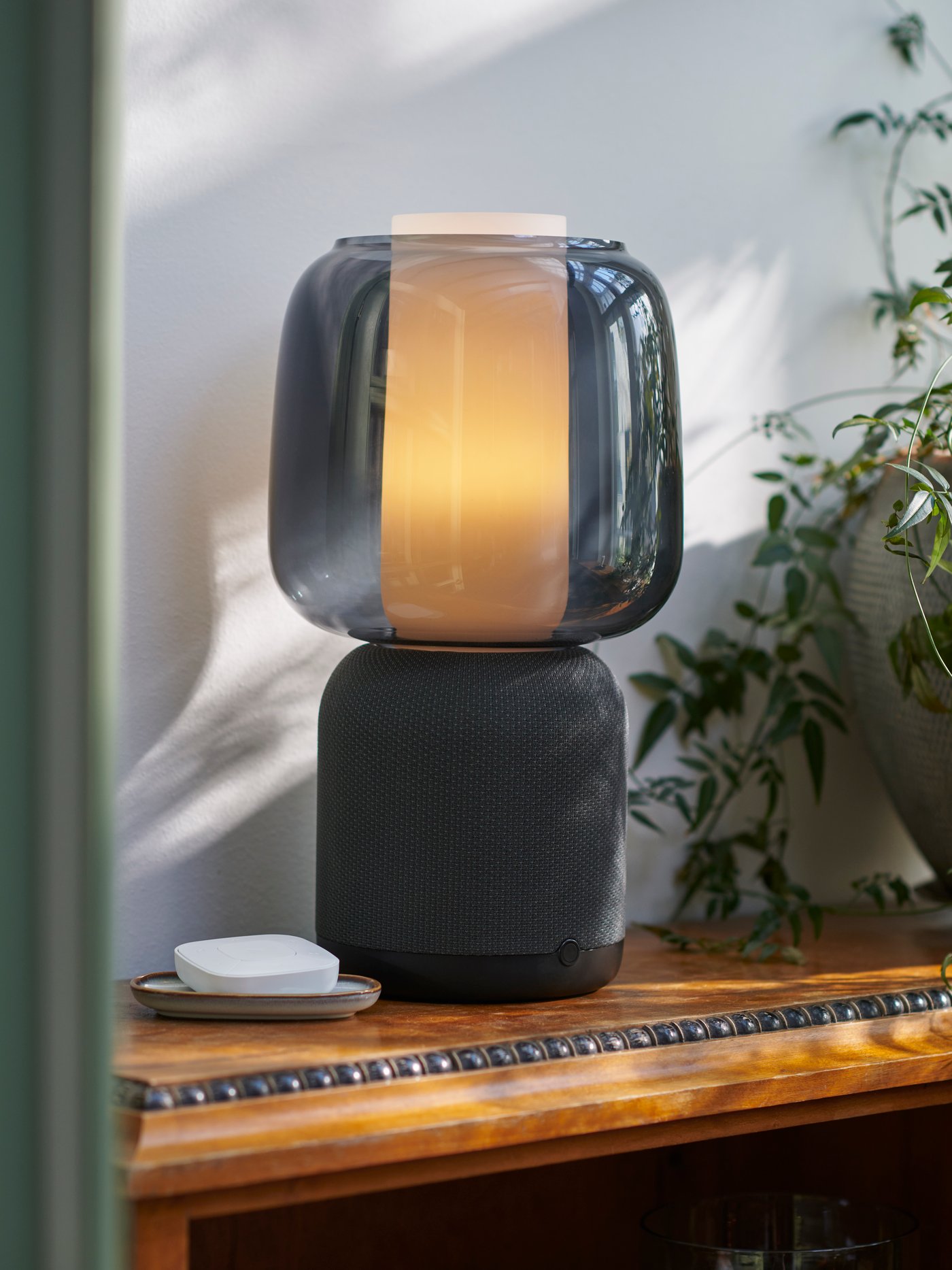 Un pied de lampe enceinte WiFi SYMFONISK noir avec un abat-jour en verre noir sur une table en bois à côté d’une plante verte.