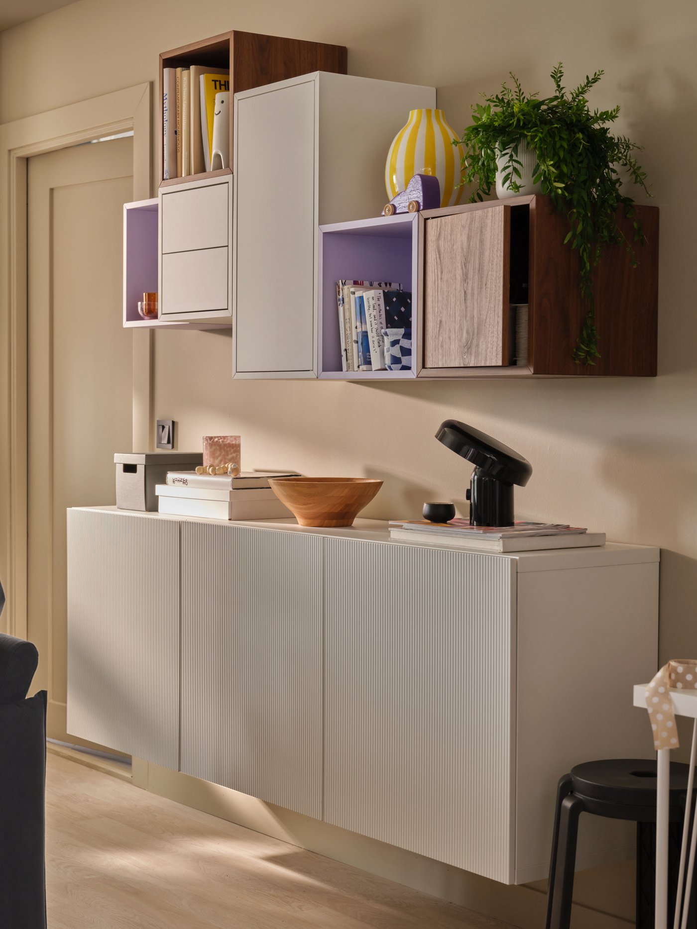 W oświetlonym łagodnymi promieniami słońca pokoju dziennym znajduje się asymetryczna kombinacja szafek wiszących EKET i BESTÅ, w których umieszczono książki i dekoracje.