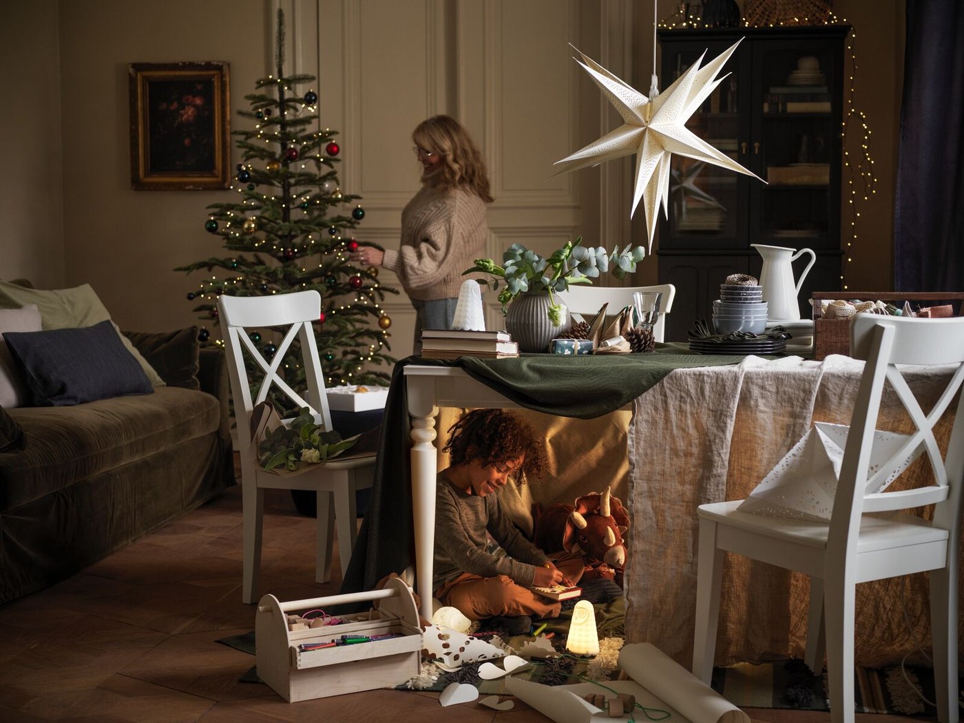 Una donna decora un albero di Natale mentre un bambino gioca sotto il tavolo, semicoperto con dei tessili per creare un nascondiglio.